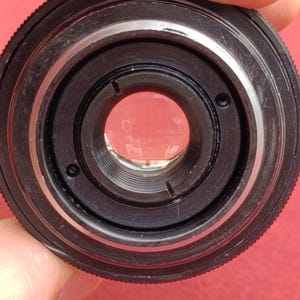 May include: Close-up of a black camera lens with silver accents. The lens has multiple concentric rings and a clear glass element in the center. The lens is held against a red background.