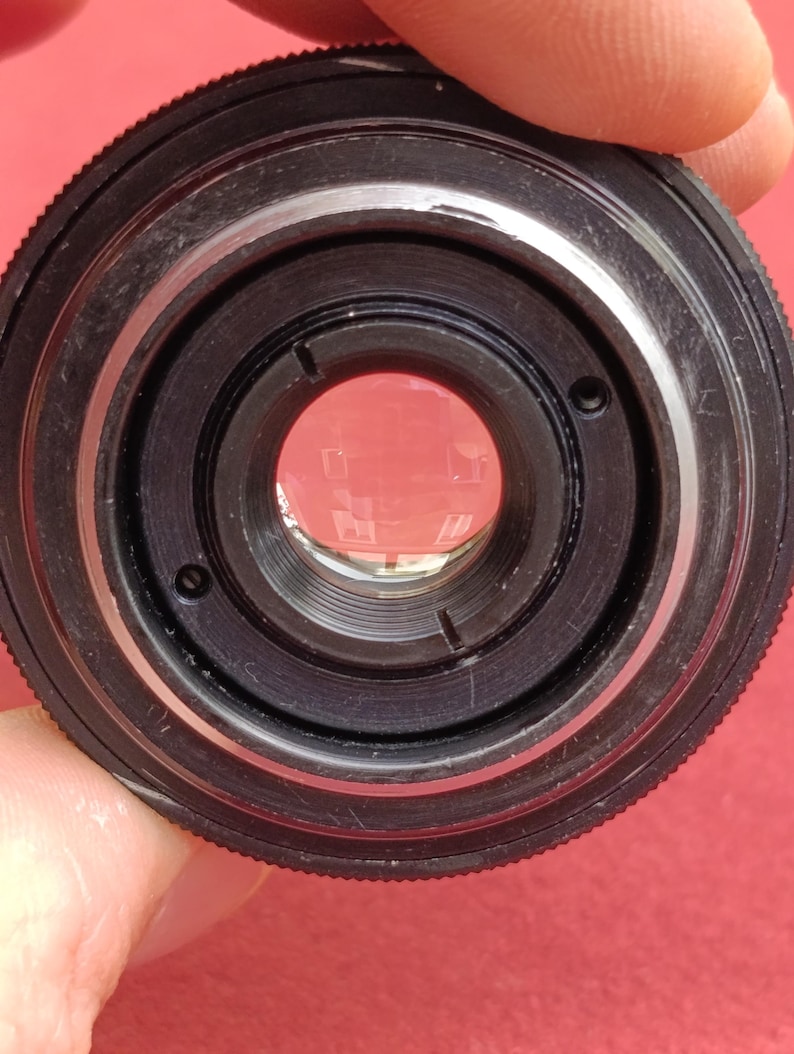 May include: Close-up of a black camera lens with silver accents. The lens has multiple concentric rings and a clear glass element in the center. The lens is held against a red background.
