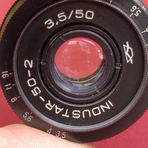 May include: Close-up of a black Industar-50-2 camera lens with the text "3.5/50". The lens has a red reflective interior and white markings. The lens is held by a person's fingers against a red background.