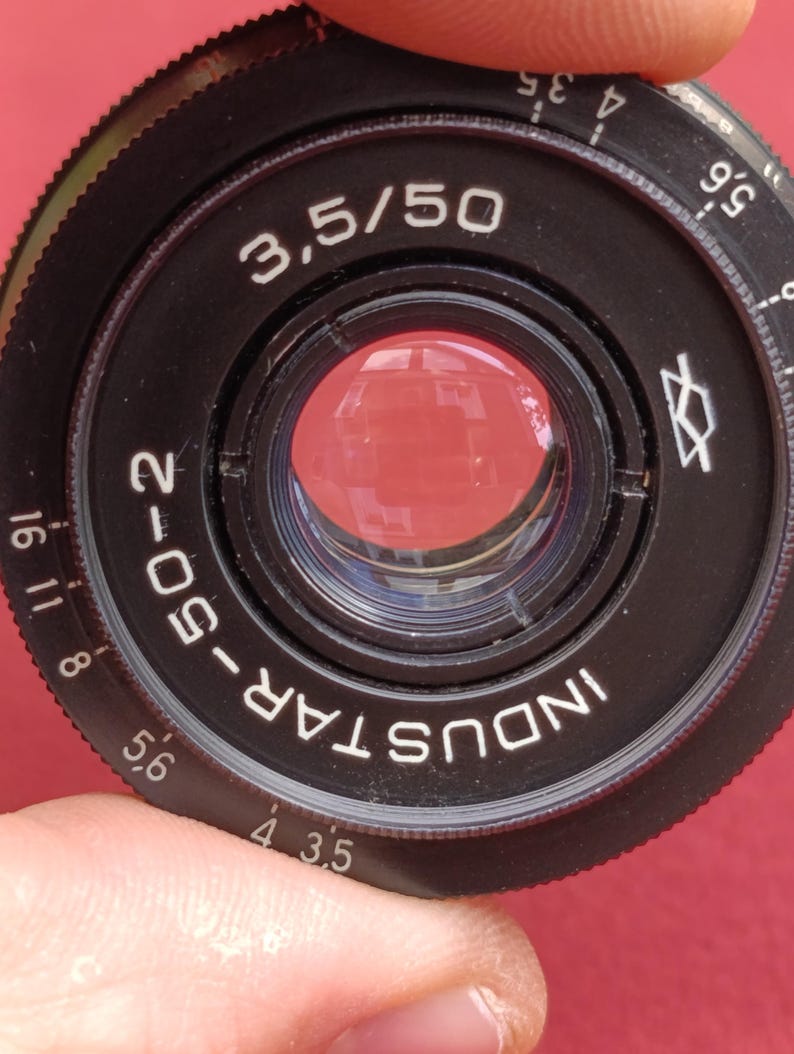 May include: Close-up of a black Industar-50-2 camera lens with the text "3.5/50". The lens has a red reflective interior and white markings. The lens is held by a person's fingers against a red background.