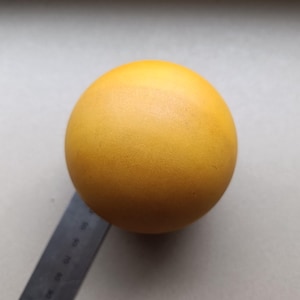Puede incluir: Una esfera amarilla sólida y lisa colocada junto a una regla negra. La regla muestra que el objeto tiene un diámetro de aproximadamente 9 cm. La esfera tiene un acabado mate y un color amarillo vibrante y uniforme en toda su superficie.