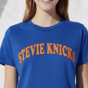 May include: A royal blue short-sleeved t-shirt featuring the text STEVIE KNICKS printed in a bold, arched orange font across the chest. The garment has a classic crew neckline and a smooth, solid-coloured fabric construction throughout.