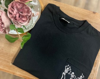 May include: Black t-shirt with a pocket displaying a white graphic of three dancing skeletons. The shirt is folded on a wooden surface, with a vase of pink roses in the background. A casual wear item.