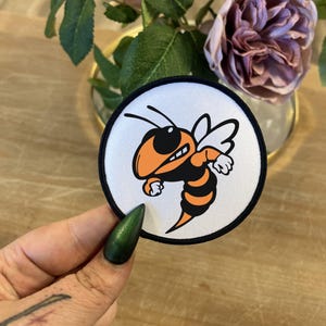 May include: A white, round patch with a black border. The patch features a cartoon image of a yellow and black bee with orange wings and a menacing expression. The bee is flying with its wings spread out.