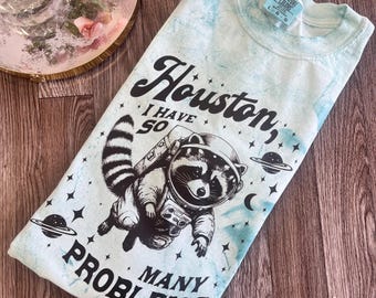 Puede incluir: Camiseta azul claro teñida con un diseño de un mapache astronauta, estrellas, planetas y la frase "Houston, I have so many problems". La camiseta está doblada sobre una superficie de madera.
