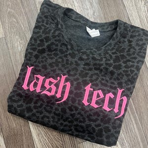 May include: Dark gray t-shirt with a leopard print pattern. The words "lash tech" are printed in bright pink, gothic-style lettering across the front. The shirt is folded on a wooden surface.