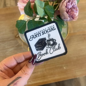 May include: A white patch with black text and a black and white illustration of a book and a cup of coffee. The text reads "Members Only Anti Social Book Club".