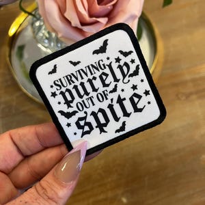 May include: White iron-on patch with black text and bats that reads "SURVIVING purely OUT OF spite".