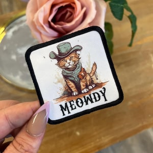 May include: A white and black embroidered patch with a cartoon cat wearing a cowboy hat and a bandana. The cat is brown and white and has a red bow tie. The text "MEOWDY" is written in black below the cat.