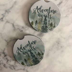 May include: Two round, white coasters with a watercolor design of a forest with the text "Adventure awaits" in black cursive lettering.