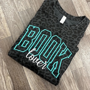 May include: A dark grey t-shirt with a leopard print pattern. The shirt has the words "BOOK lover" in a teal and white design. The t-shirt is folded and placed on a wooden surface.