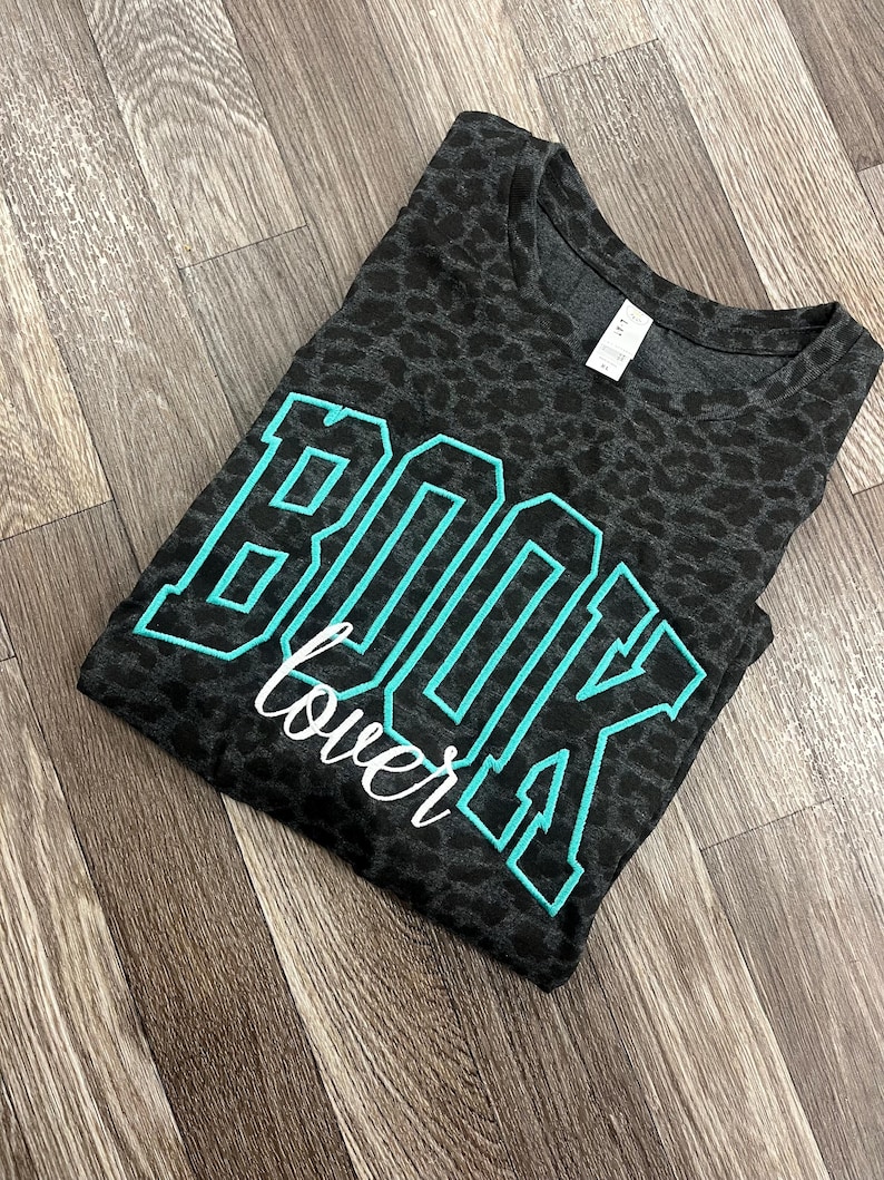 May include: A dark grey t-shirt with a leopard print pattern. The shirt has the words "BOOK lover" in a teal and white design. The t-shirt is folded and placed on a wooden surface.