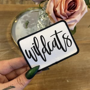May include: White fabric patch with black embroidered text that reads "Wildcats".