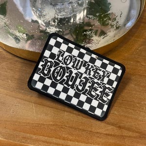 May include: Black and white checkered patch with the text "Low Key Boujee" in a black gothic font.