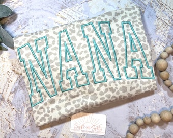 May include: A white t-shirt with a light gray leopard print pattern. The word "NANA" is embroidered in teal blue on the shirt.