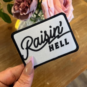 May include: White iron-on patch with black text that reads "Raisin' Hell".