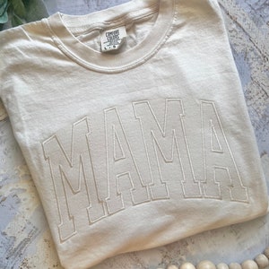 May include: A beige t-shirt with the word "MAMA" embroidered in white on the front. The shirt is made by Comfort Colors and is size L/G.