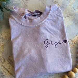 May include: A lavender-colored t-shirt with the embroidered word "Gigi" and a small purple heart. The shirt is folded, showing the neckline and the embroidered design on the upper left chest area. The fabric appears soft and comfortable.