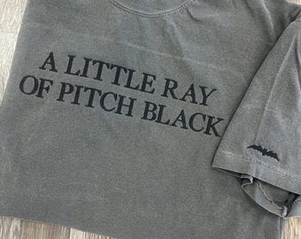 May include: A gray t-shirt with the text "A LITTLE RAY OF PITCH BLACK" in black. The sleeve has a small embroidered bat design. The shirt is made of a soft, worn material.