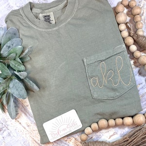 May include: A sage green t-shirt with a pocket on the front. The pocket has the letters "akl" embroidered on it in a gold thread. The t-shirt is made by "By Miss Sally Handmade".