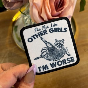May include: Square patch with a black border and the text "I'm Not Like Other Girls I'm Worse." The patch features a cartoon raccoon illustration in black and white. The patch is held up by a hand.