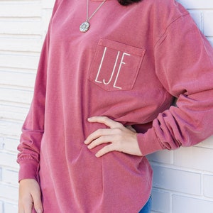 May include: A long-sleeved, maroon-colored shirt with a pocket on the chest. The pocket features the embroidered initials "LJE" in white. The shirt is worn casually, with the person leaning against a white brick wall.