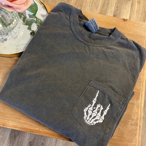 K&ouml;nnte beinhalten: Ein dunkelgraues Langarmshirt mit einer wei&szlig;en Skeletthand, die das Rock'n'Roll-Zeichen in einer Tasche auf der Vorderseite des Shirts macht.