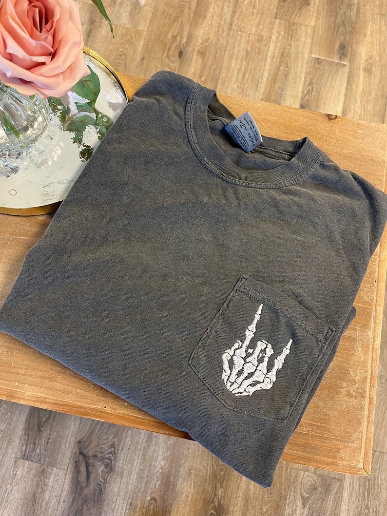 K&ouml;nnte beinhalten: Ein dunkelgraues Langarmshirt mit einer wei&szlig;en Skeletthand, die das Rock'n'Roll-Zeichen in einer Tasche auf der Vorderseite des Shirts macht.