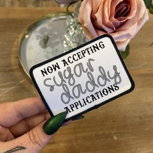 May include: A white rectangular patch with a black border, featuring the text "NOW ACCEPTING SUGAR DADDY APPLICATIONS" in a decorative font. The patch is held in front of a blurred background with flowers and a mirror.