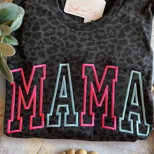 May include: A dark gray t-shirt with a leopard print pattern. The word "MAMA" is embroidered across the front in pink with a light blue outline. A tag that says "By Miss Sally Handmade" is attached to the shirt.