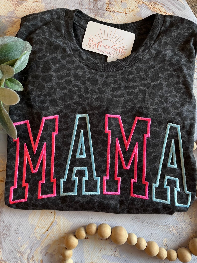 May include: A dark gray t-shirt with a leopard print pattern. The word "MAMA" is embroidered across the front in pink with a light blue outline. A tag that says "By Miss Sally Handmade" is attached to the shirt.