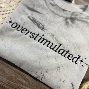May include: A light gray t-shirt with the word "overstimulated" printed in black script. The shirt has a distressed, vintage look with star accents. The fabric appears soft and slightly textured.