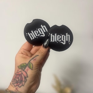 May include: Two black car coasters with the word "blegh" in white gothic lettering. The coasters are round with a small notch. The background is a plain white wall.