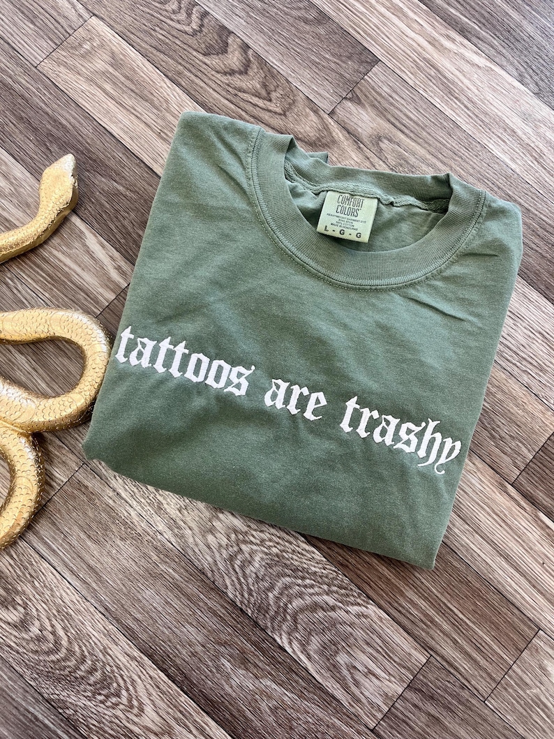 May include: An olive green Comfort Colors t-shirt with the text "tattoos are trashy" in white gothic lettering. The shirt is folded and lies on a wooden surface. A gold-colored snake figurine is in the upper left corner.