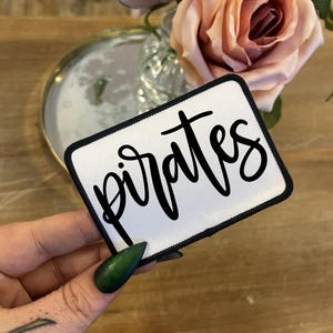May include: A rectangular white patch with the word "pirates" in black script lettering. The patch has a black border. The background includes a wooden surface, a mirror, and pink roses.