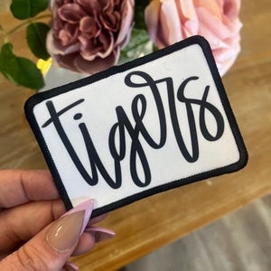 May include: White iron-on patch with black cursive text that says "Tigers". The patch has a black border.