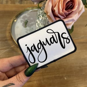 May include: A white rectangular patch with the word "jaguars" in black cursive script. The patch has a black border. The background includes a wooden surface, a mirror, and pink roses.