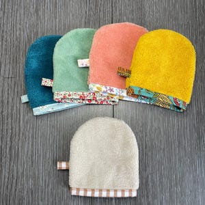 May include: Five rounded terry cloth mitts in teal, sage green, coral, mustard yellow, and beige. Each mitt features a small fabric loop on the side and a decorative patterned fabric trim along the bottom edge, including floral and gingham prints.