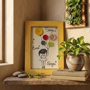Op de afbeelding: Een ingelijst kunstwerk met een gele rand toont een tekening van een meisje dat knoopballonnen vasthoudt. Het kunstwerk bevat de tekst "Segui i tuoi Sogni". Een kleine vetplant is aan de muur bevestigd.