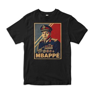 Può includere: Una t-shirt nera a maniche corte con una stampa grafica di una persona in uniforme militare con medaglie e berretto. Lo sfondo della stampa è diviso in sezioni diagonali blu e rosse. Il nome MBAPPÉ è stampato in grassetto color beige.
