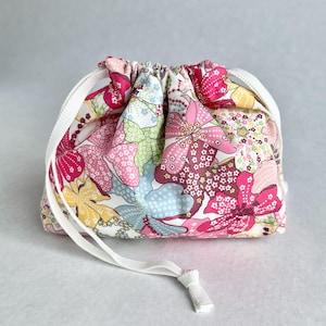 May include: A fabric drawstring pouch featuring a vibrant floral pattern with shades of pink, yellow, light blue, and green on a white background. The bag is cinched at the top with a white woven ribbon drawstring that hangs down the front.