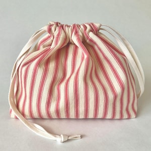 May include: A small drawstring bag with a red and cream striped pattern. The bag is made of fabric and has a cream-colored drawstring closure. The bag is designed to hold small items.