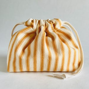 Könnte beinhalten: Ein Kordelzugbeutel mit vertikalen Streifen in Creme und Goldgelb. Der Beutel hat eine abgerundete Form mit geraffter Oberseite und Kordelzugverschluss. Die Griffe und der Kordelzug sind einfarbig cremefarben.