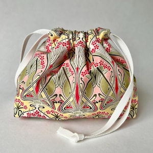 Może przedstawiać: Worek ze sznurkiem ze wzorem kwiatowym w kolorze żółtym, różowym, czerwonym i zielonym. Torba ma białe wstążki i białe zapięcie na wstążkę. Torba jest prawdopodobnie wykonana z tkaniny.