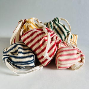 May include: A collection of small fabric drawstring pouches featuring vertical stripes in various colors including red, navy blue, dark green, yellow, and pink on an off-white background. Each pouch has a matching off-white cotton drawstring cord.