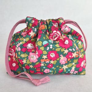 Puede incluir: Una bolsa con cordón que presenta un vibrante estampado de flores y estrellas sobre un fondo verde oscuro. La tela está decorada con flores rosas, rojas y naranjas intercaladas con pequeñas estrellas beige. Incluye dos cordones de cinta rosa.
