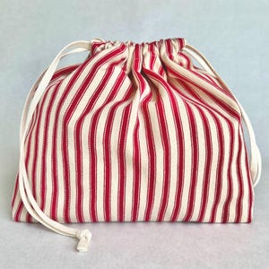 May include: A drawstring pouch featuring a vertical striped pattern in red and off-white. The bag is constructed from a woven fabric and includes two integrated off-white fabric cords that cinch the top opening closed, creating a gathered effect.