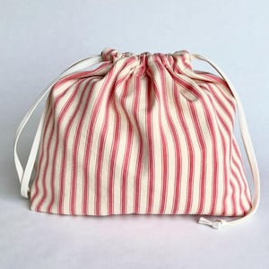 May include: A drawstring pouch featuring a vertical striped pattern in red and off-white. The bag is constructed from a woven fabric and includes two white cord drawstrings that gather the top opening, creating a soft, pleated texture.