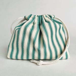 May include: A small drawstring bag featuring teal and off-white vertical stripes. The bag is made of fabric and has a white drawstring closure. The bag is designed for carrying small items and has a simple, classic design.