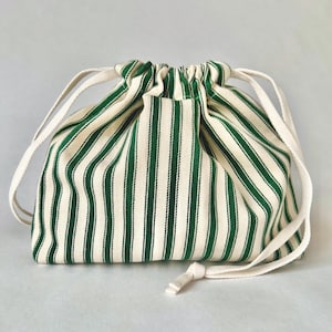 May include: A fabric drawstring bag featuring a cream background with vertical green stripes. It has a gathered top and a white drawstring closure. The bag is designed for carrying small items and is made of fabric.